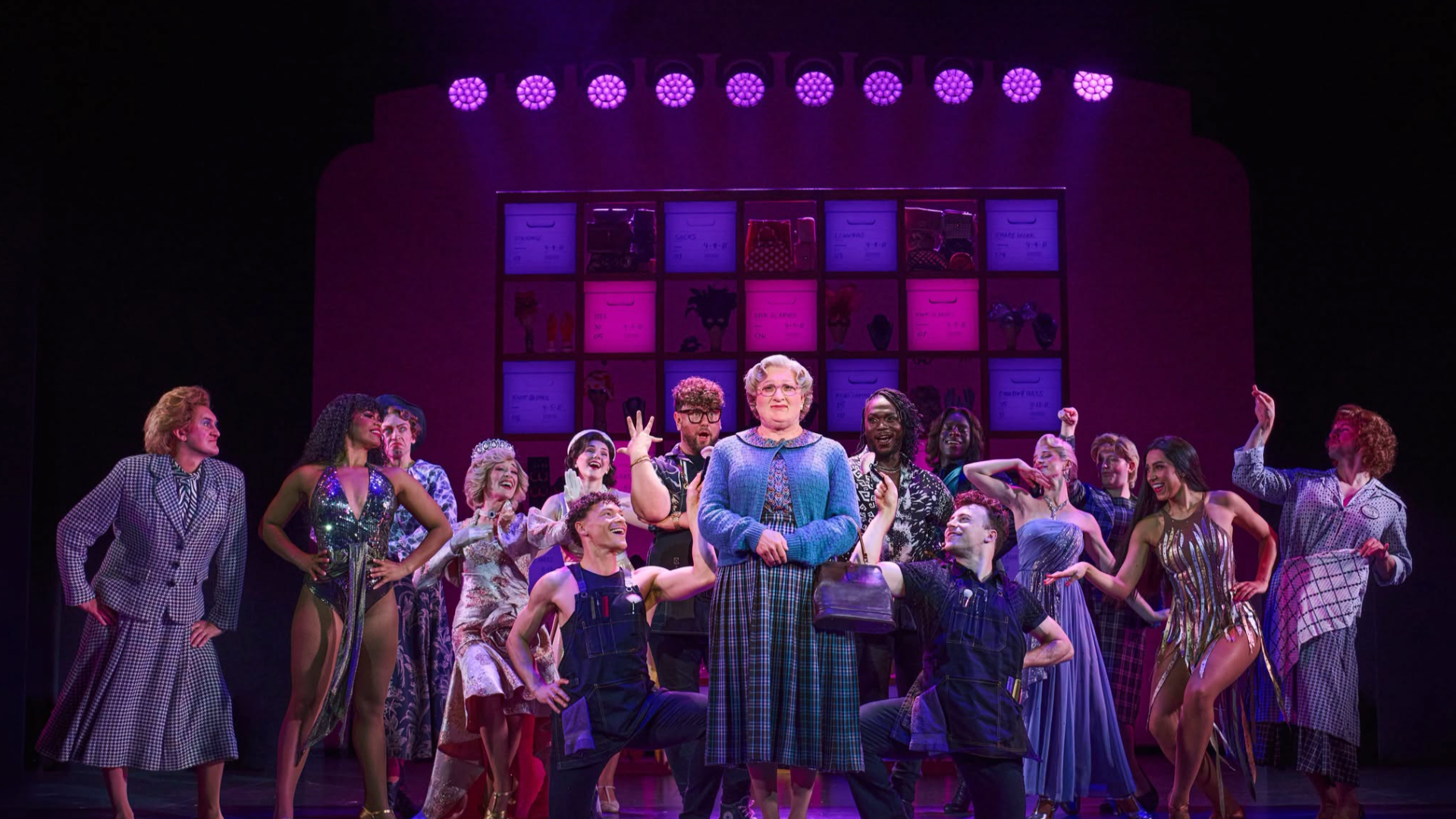 Mrs. Doubtfire stands centre stage surrounded by the ensemble under vivid purple lighting.
