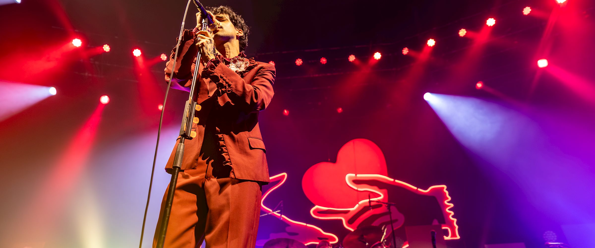 Stephen Sanchez performs on stage during his Love Sings Again European Tour, singing into a microphone beneath vibrant red, purple and white stage lighting. Multiple lighting fixtures and beams are visible above and around the stage, while a neon heart backdrop glows behind the band.