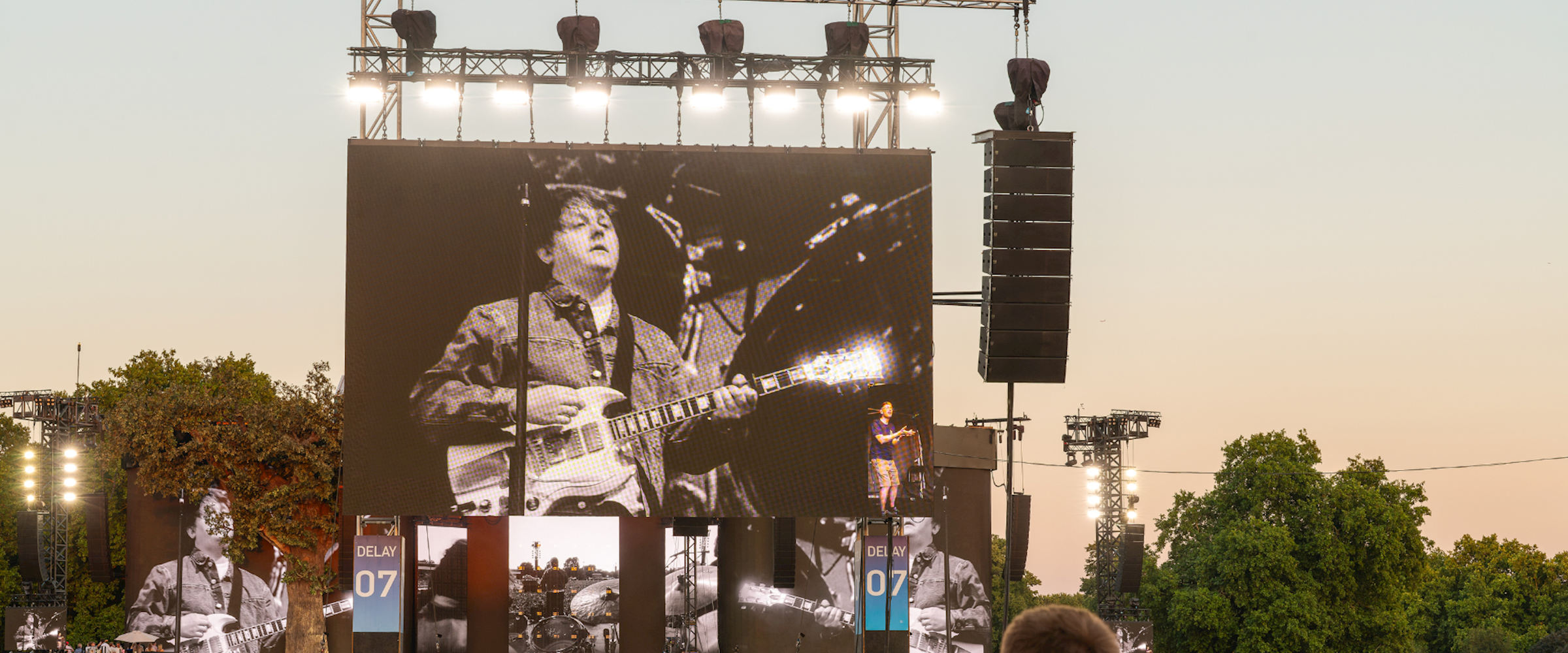 Lewis Capaldi performing on the Great Oak Stage at BST Hyde Park, displayed on a large LED screen above the stage, with thousands of festivalgoers watching from the audience as the sun sets. PRG's video screens, audio arrays and stage infrastructure are visible throughout the scene.