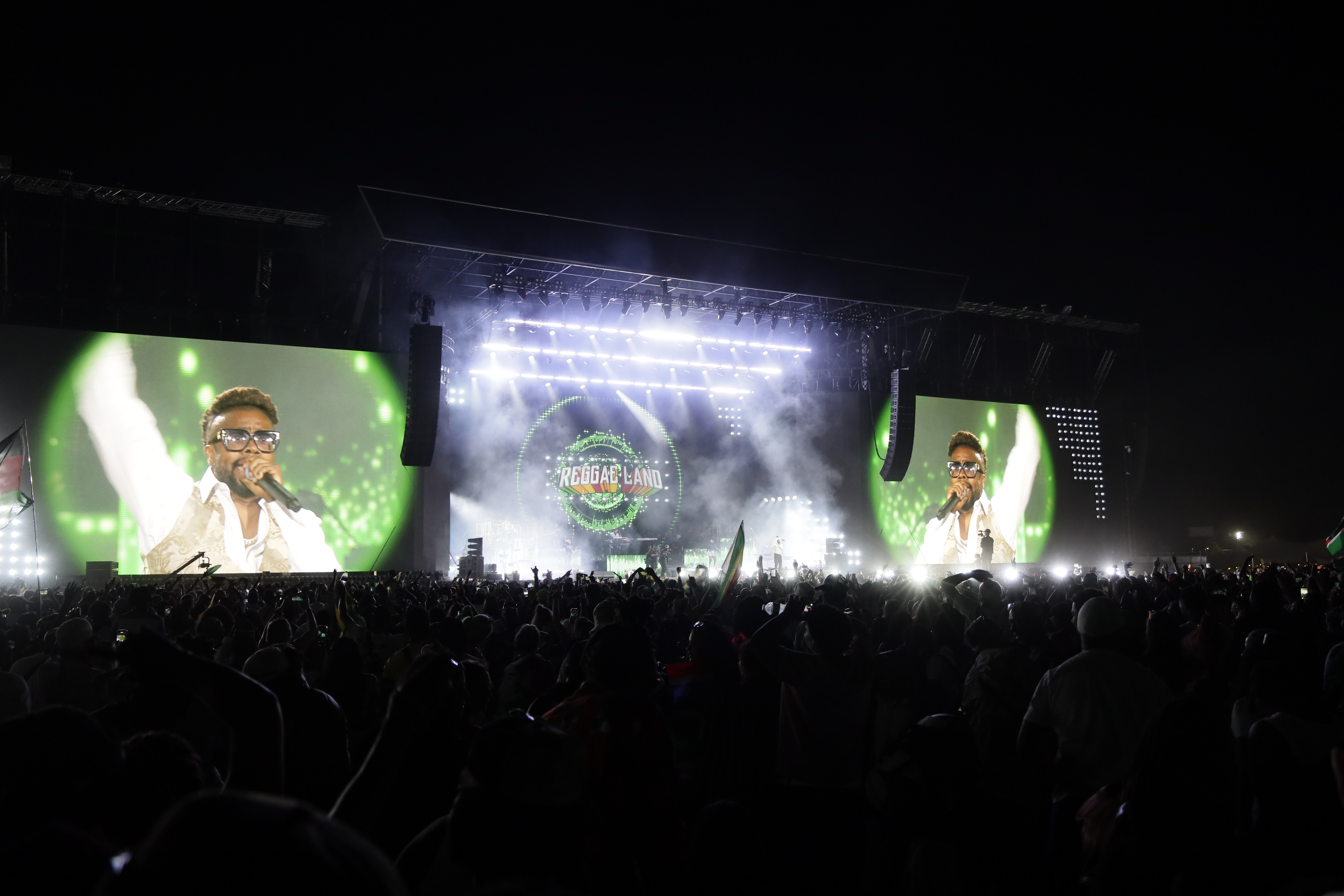 A performer in white sings into a microphone on two large screens at Reggae Land, as bright lights cut through stage smoke above the crowd.