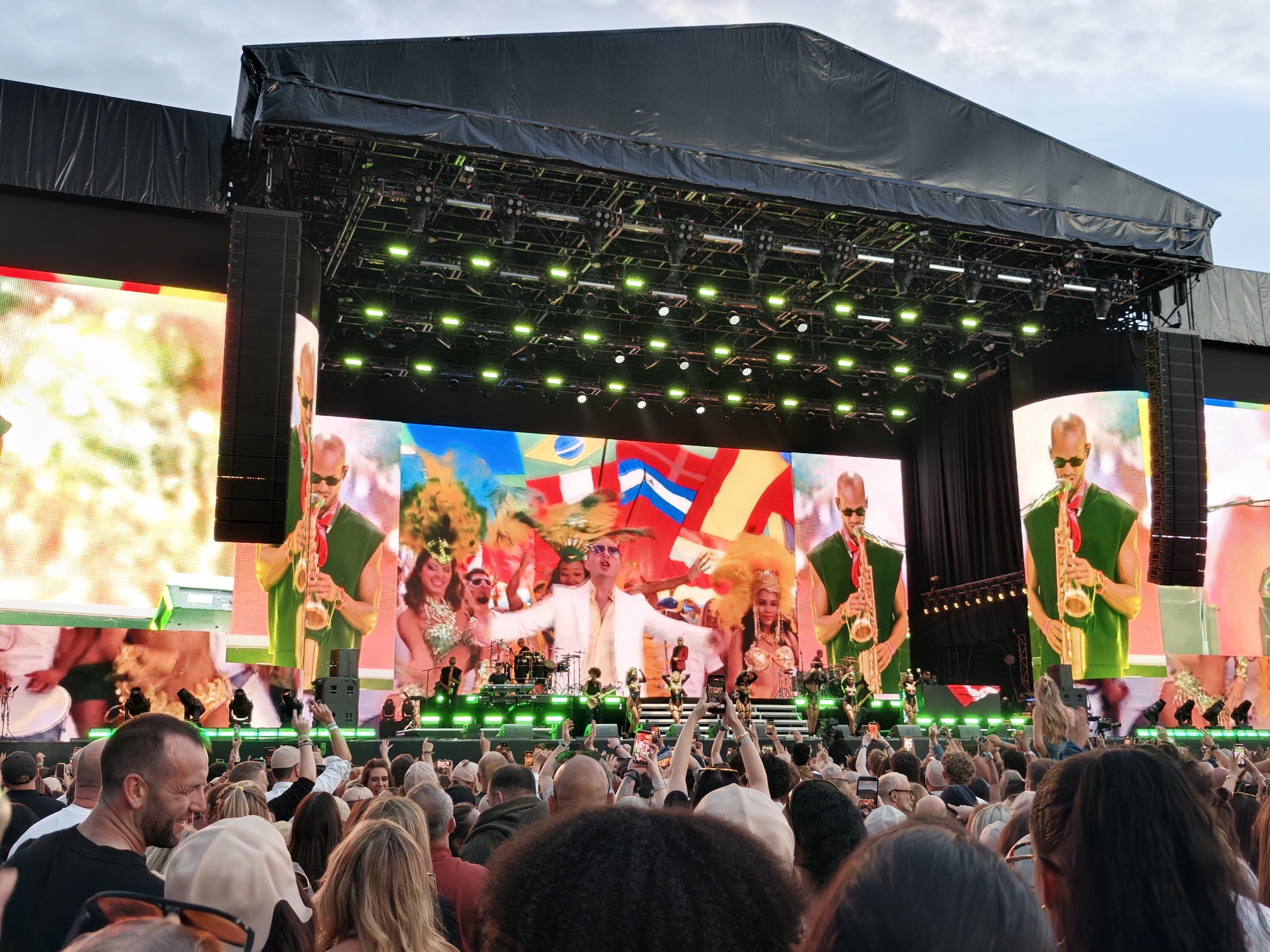 Pitbull performs on the main stage at the inaugural Roundhay Festival in Leeds, with vibrant LED screens, green stage lighting and a packed crowd watching from the audience.