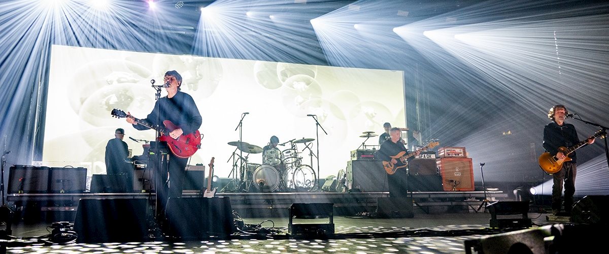 Super Furry Animals perform live at the O2 Apollo Manchester beneath dramatic white concert lighting that fills the stage with sweeping beams. The band members are positioned across the stage with guitars, drums and keyboards, while a wide LED screen displays a soft abstract backdrop. Circular lighting patterns cover the stage floor, creating an atmospheric live performance scene.