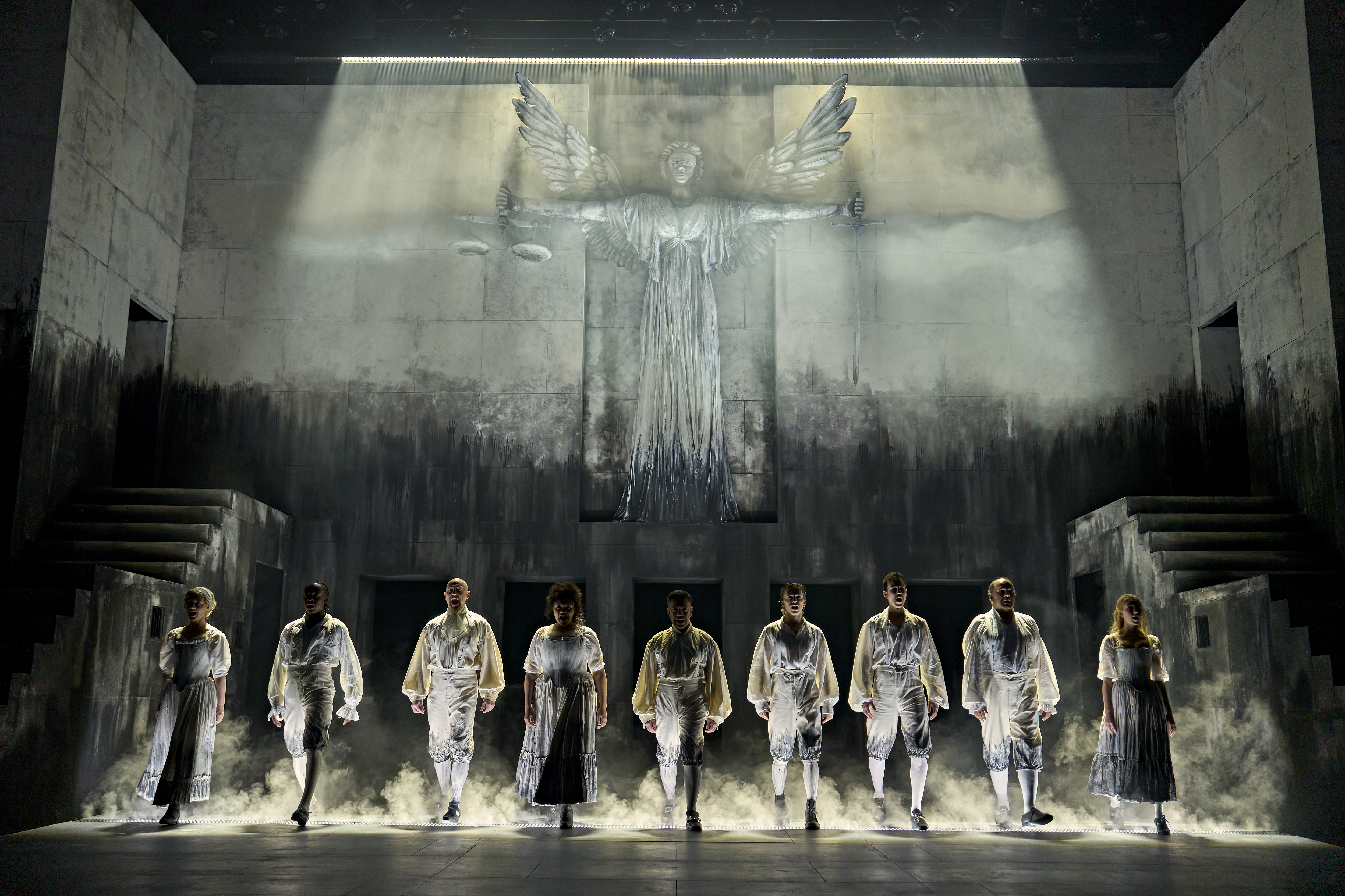 The cast of Sweeney Todd performs beneath a dramatic winged statue on the Birmingham Rep stage, with atmospheric lighting, haze and monochrome scenery creating the production's dark and haunting visual aesthetic.